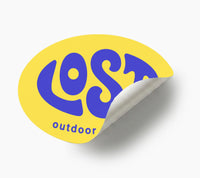A yellow oval peel-and-stick sticker with the words 'LOST outdoor' in bold blue lettering, shown on a light gray background with a curled bottom-right corner revealing the backing.