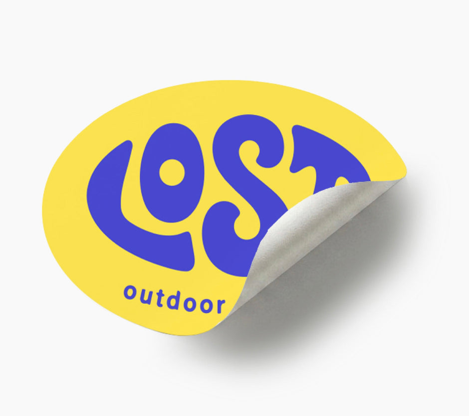 A yellow oval peel-and-stick sticker with the words 'LOST outdoor' in bold blue lettering, shown on a light gray background with a curled bottom-right corner revealing the backing.